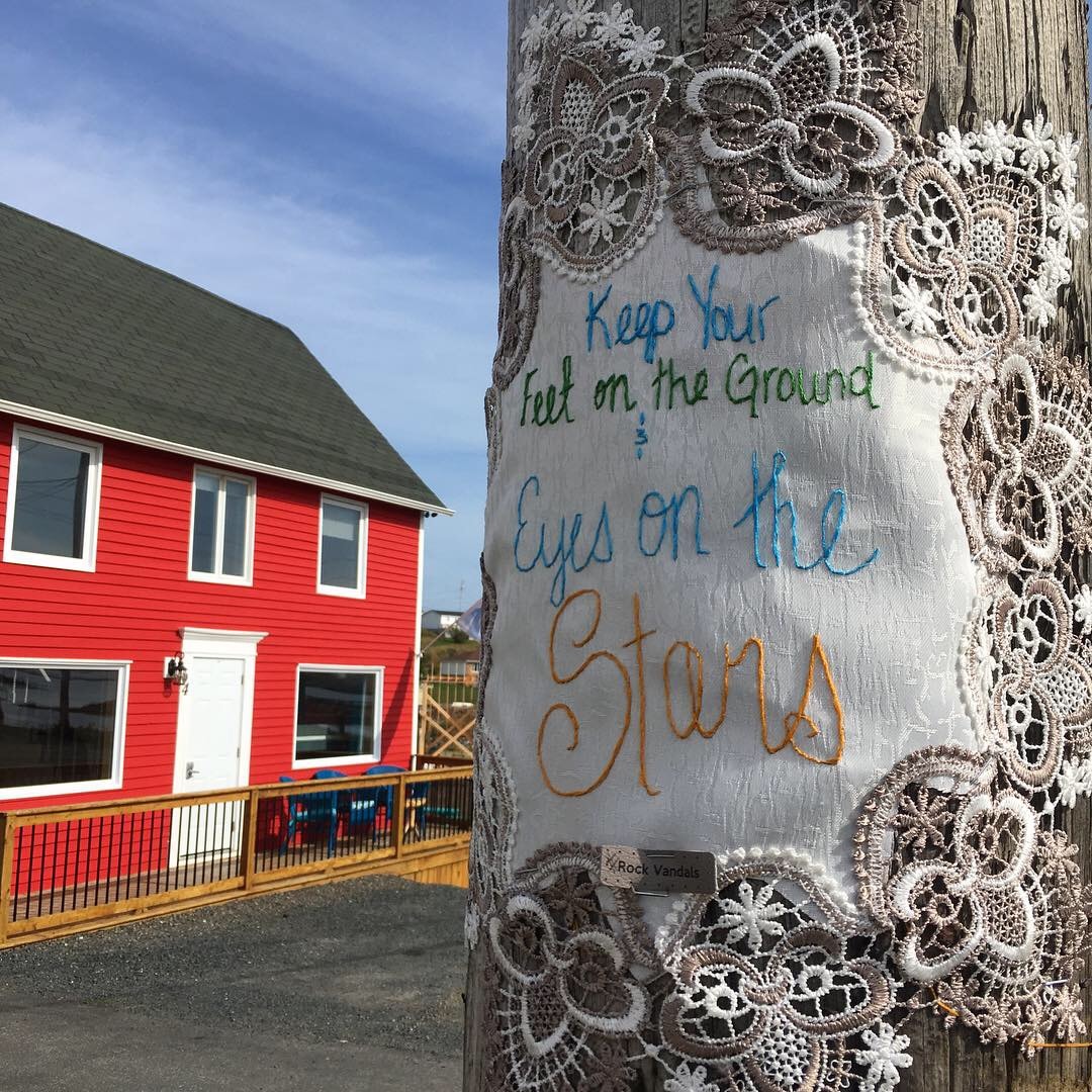 A colorful red building stands in the background, complemented by a lace-adorned cloth affixed to a wooden pole. The cloth features handwritten text encouraging focus on both the ground and the stars, enhancing the local atmosphere.
