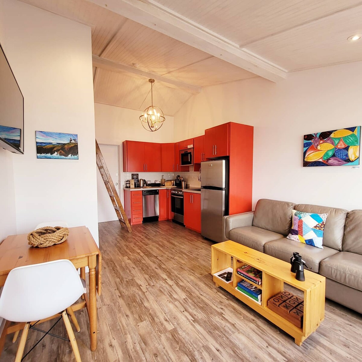 An open concept kitchen and living area features bold red cabinetry and stainless-steel appliances. A cozy seating arrangement includes a grey sofa with a colorful accent pillow. A wooden table adds warmth, while local artwork adorns the walls, enhancing the inviting space.