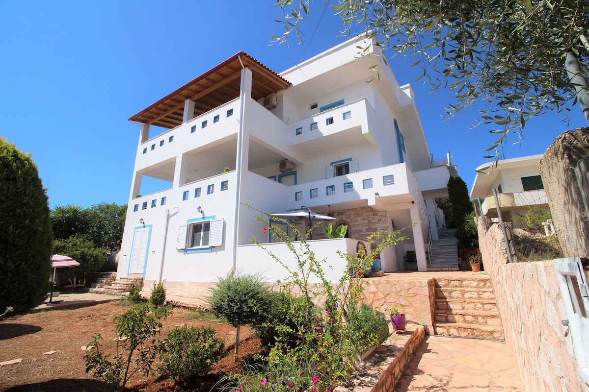 A two-story building is shown, featuring a white exterior and multiple balconies. The surrounding garden is landscaped with greenery and flower pots, providing a serene setting. A staircase leads to the entrance, which is visible from the well-maintained outdoor area.