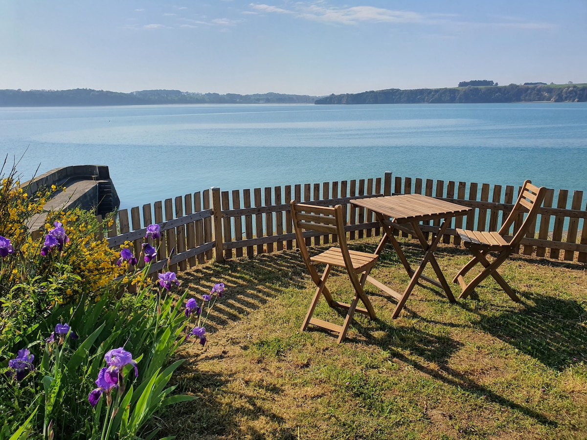 A scenic outdoor area features a small wooden table and two folding chairs, surrounded by green grass and blooming purple irises. A wooden fence encloses the space, providing a picturesque view of the calm, blue water and distant cliffs under a clear sky.
