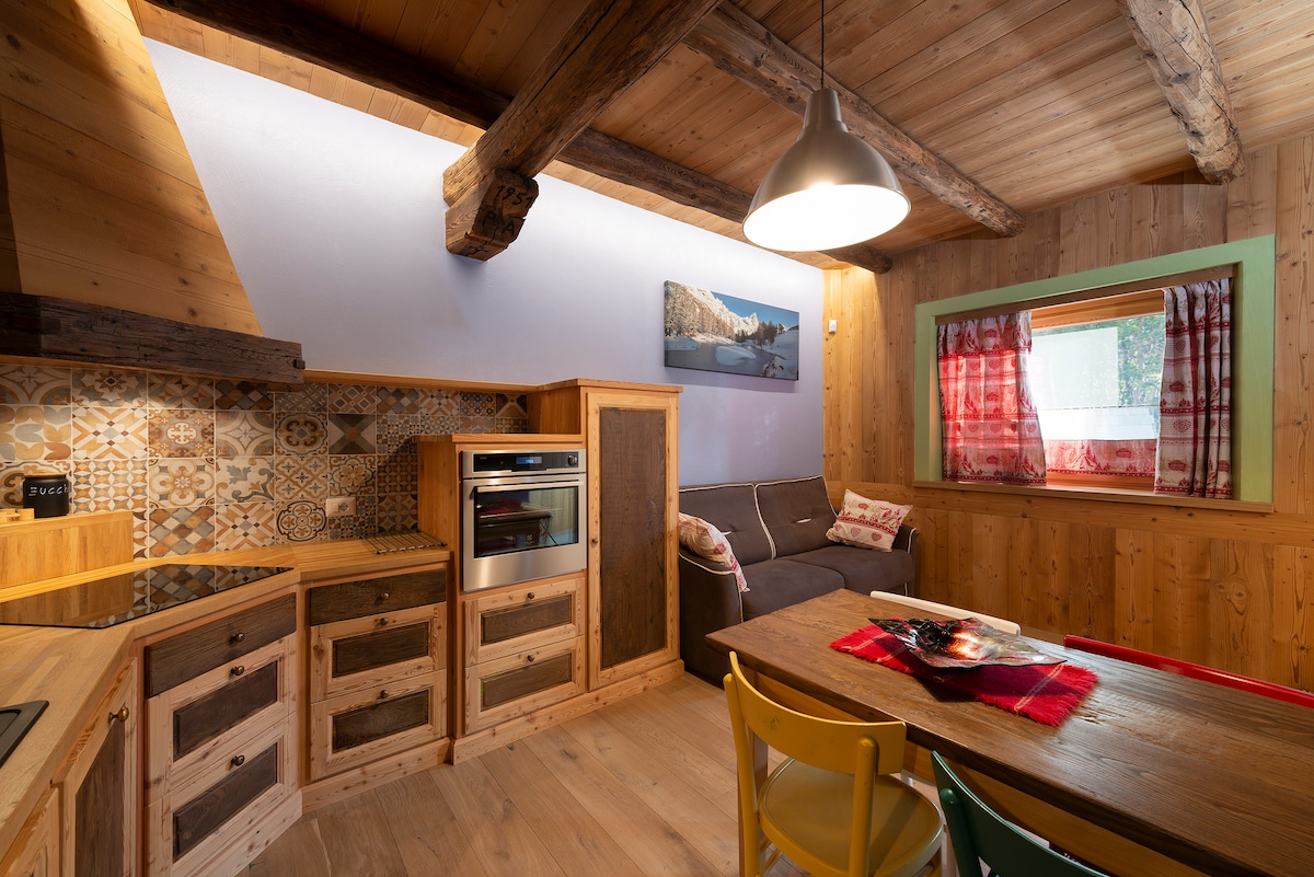 A well-appointed kitchen area showcases wooden cabinetry and a modern oven. A cozy seating space with a sofa is visible, complemented by a dining table adorned with a red tablecloth. Bright windows, framed in green, allow natural light to fill the space.