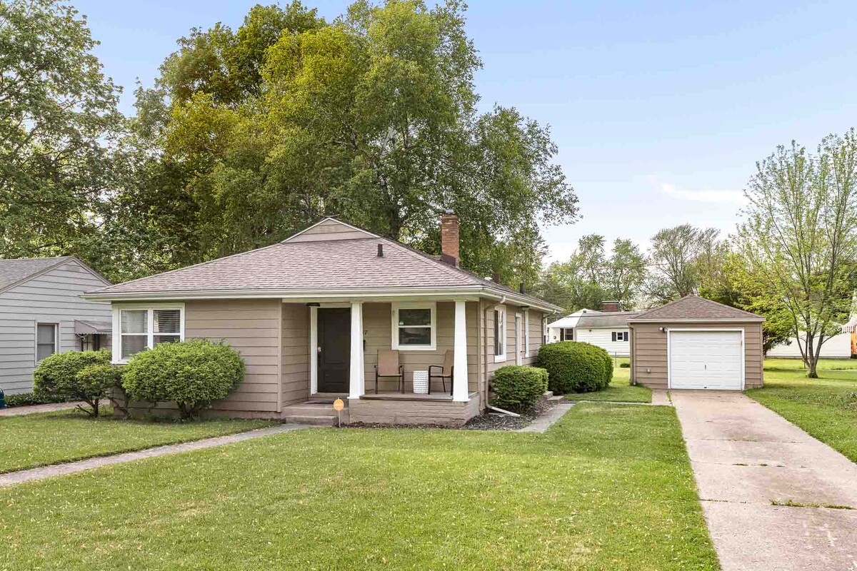 A single-family home features a welcoming front porch with two simple chairs. The well-maintained yard is surrounded by lush greenery, leading to a nearby garage. A paved driveway provides access, while a calm neighborhood ambiance is evident in the distance.