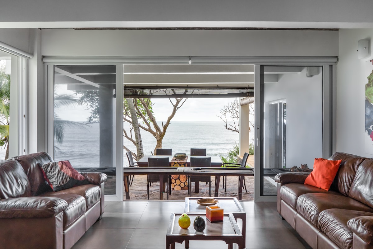 A spacious living area features comfortable leather sofas facing an opening that leads to a dining area with a view of the ocean. Large glass doors allow natural light to fill the space, with a wooden dining table set for outdoor meals against the backdrop of the sea.