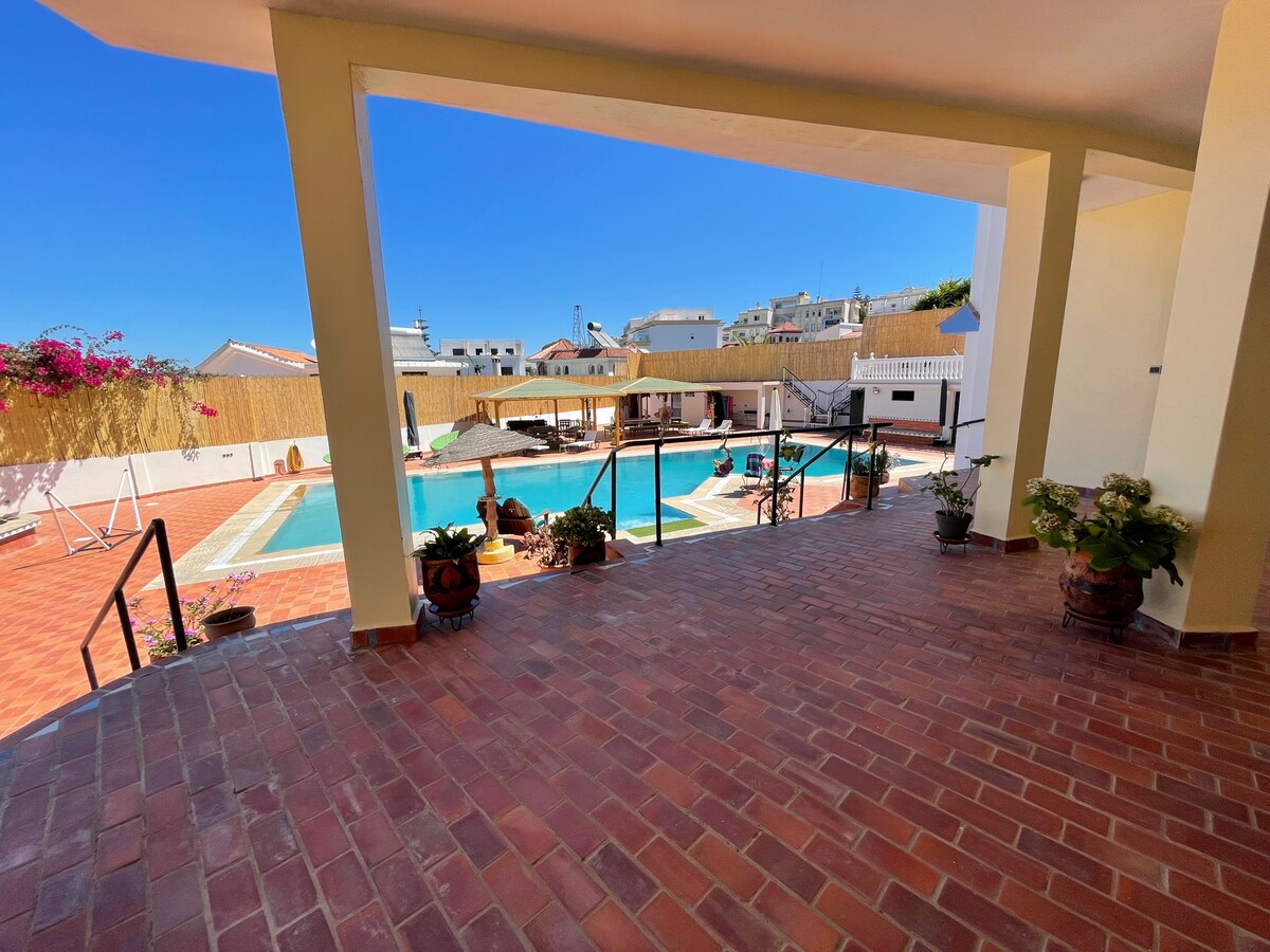 A spacious terrace with terracotta tiles overlooks the shared pool area. Lush potted plants are positioned along the railing, and a clear blue sky frames the view. The pool area features loungers and a shaded pavilion, creating an inviting environment for relaxation.