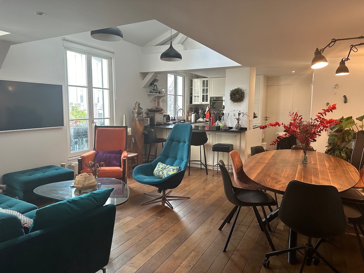 The living space features a blend of seating with vibrant blue and green chairs alongside a wooden dining table. A flat-screen television is mounted on the wall, while bright windows provide ample natural light. Decorative plants and an artistic centerpiece add character to the area.