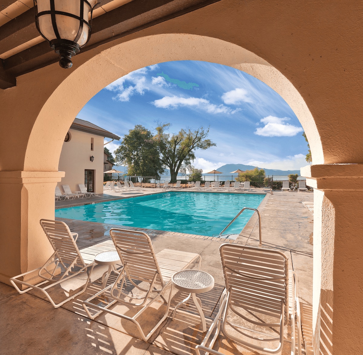An arched entrance frames a sparkling outdoor pool surrounded by lounge chairs. The scene features clear blue skies and trees in the background, creating a serene oasis for relaxation at the resort.