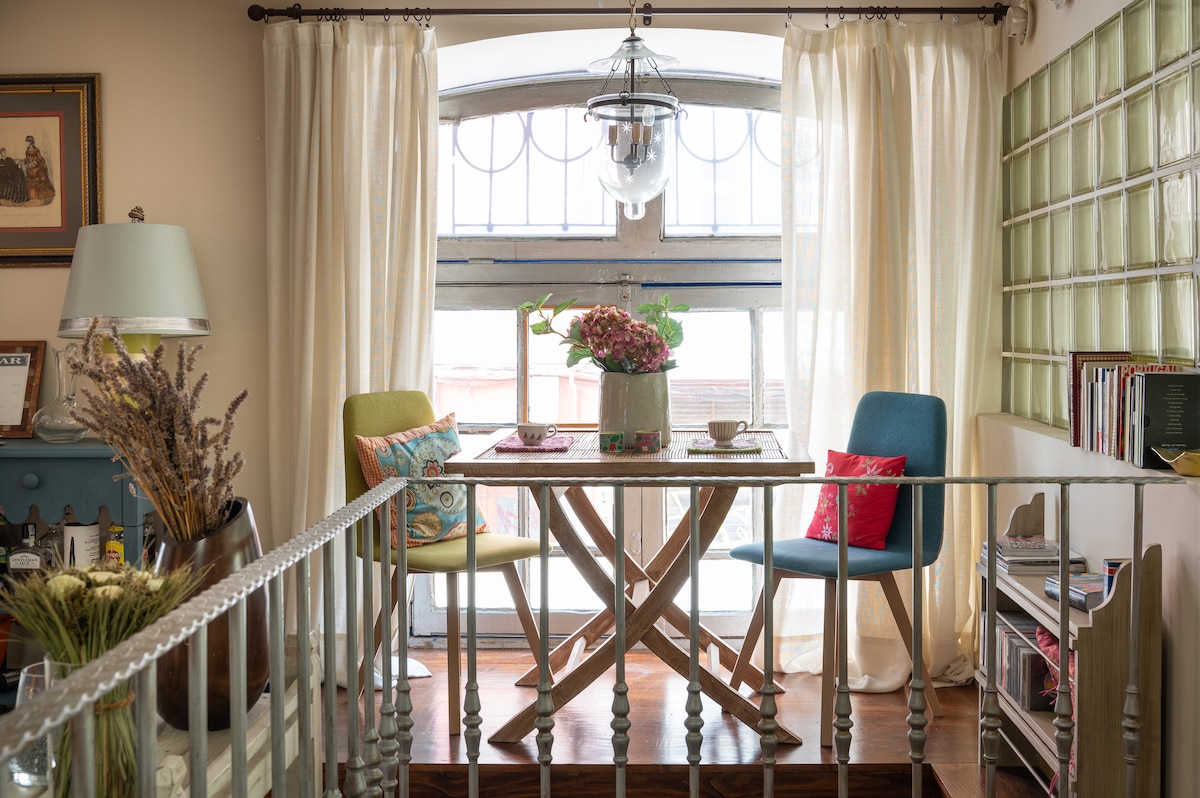 A dining area features a wooden table with two colorful chairs and a floral centerpiece. Natural light filters through large windows, complemented by flowing curtains. Decorative shelving with books and small accessories enhances the inviting space, emphasizing a blend of comfort and style.