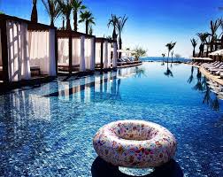 An expansive infinity pool stretches towards the horizon, bordered by elegant cabanas and palm trees. A colorful inflatable swim ring floats on the shimmering water, reflecting the clear blue sky above.