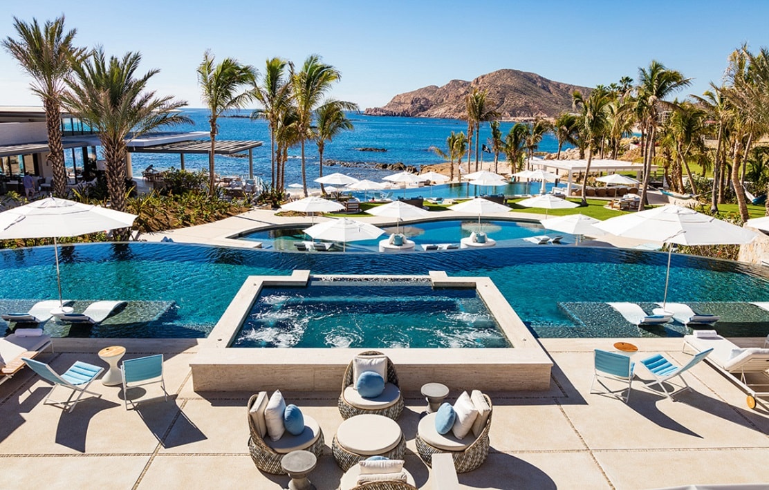 An inviting pool area is showcased, featuring multiple infinity pools bordered by palm trees. Sun loungers and umbrellas are positioned around the water, with ocean views in the background. A jacuzzi sits prominently, enhancing the resort-style atmosphere.