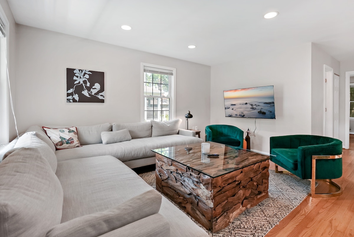 A spacious living area is furnished with a large sectional sofa and two green accent chairs. A unique wooden coffee table centers the space, complemented by a neutral area rug. Natural light enters through windows, enhancing the calm ambiance.