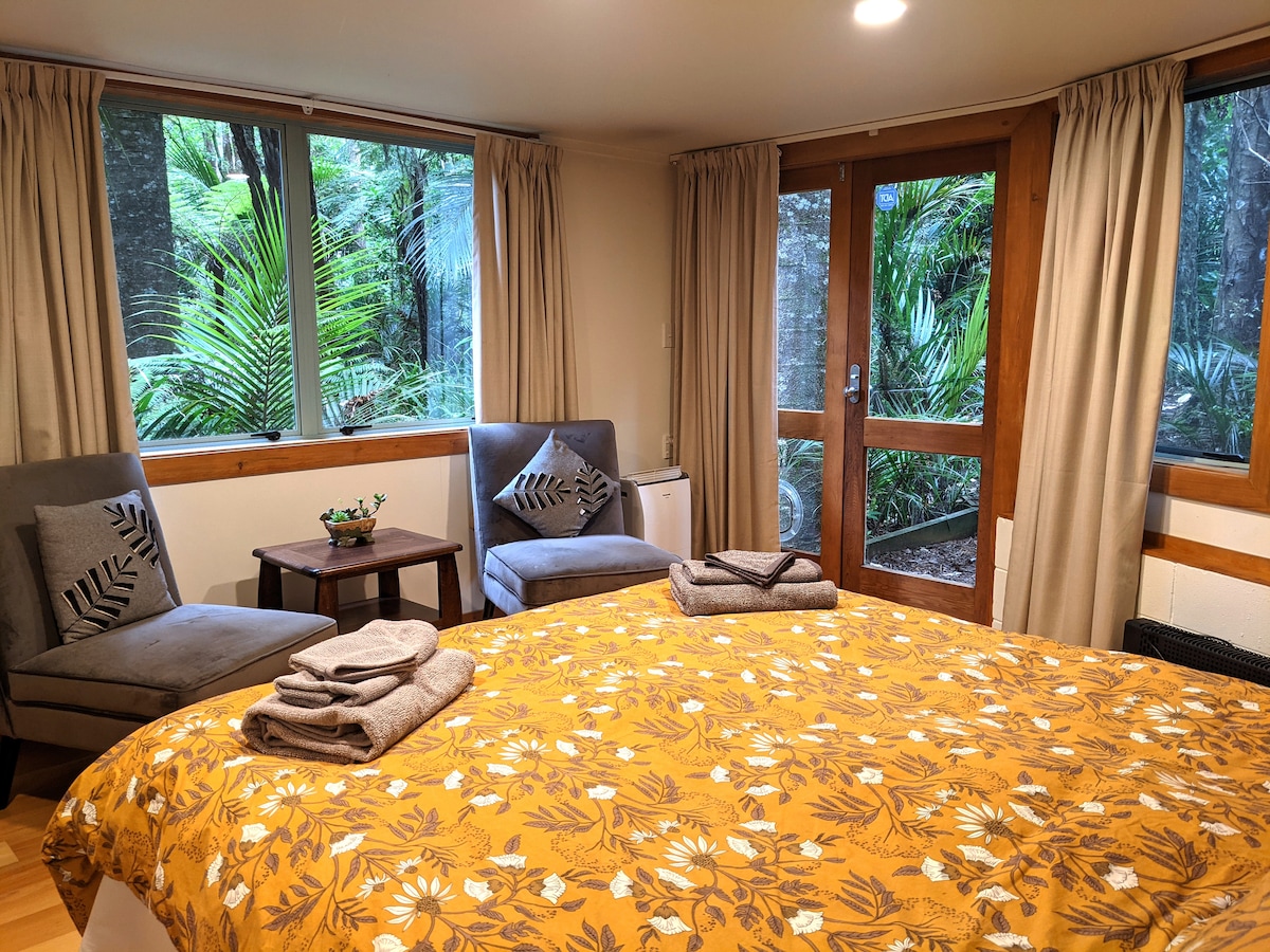 A cozy room features a spacious bed adorned with a floral-patterned duvet cover, flanked by two chairs and a small table. Large windows invite natural light and offer views of the surrounding greenery, while curtains add a touch of privacy.