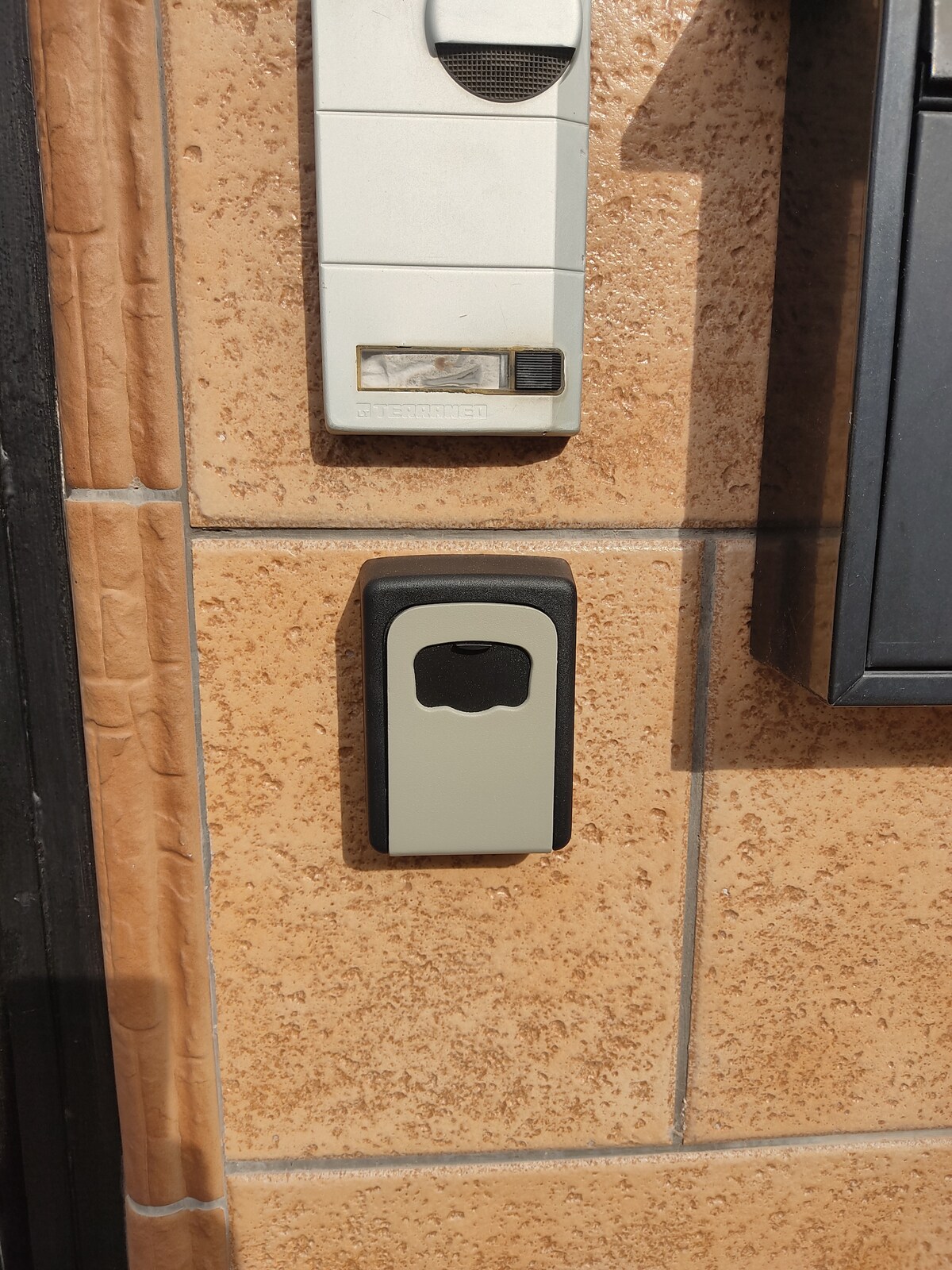 An entry panel is mounted on a textured wall, accompanied by a modern intercom system. A secure key lockbox is positioned below, providing convenient access for guests. The surface features warm, earthy tones that complement the overall aesthetic.