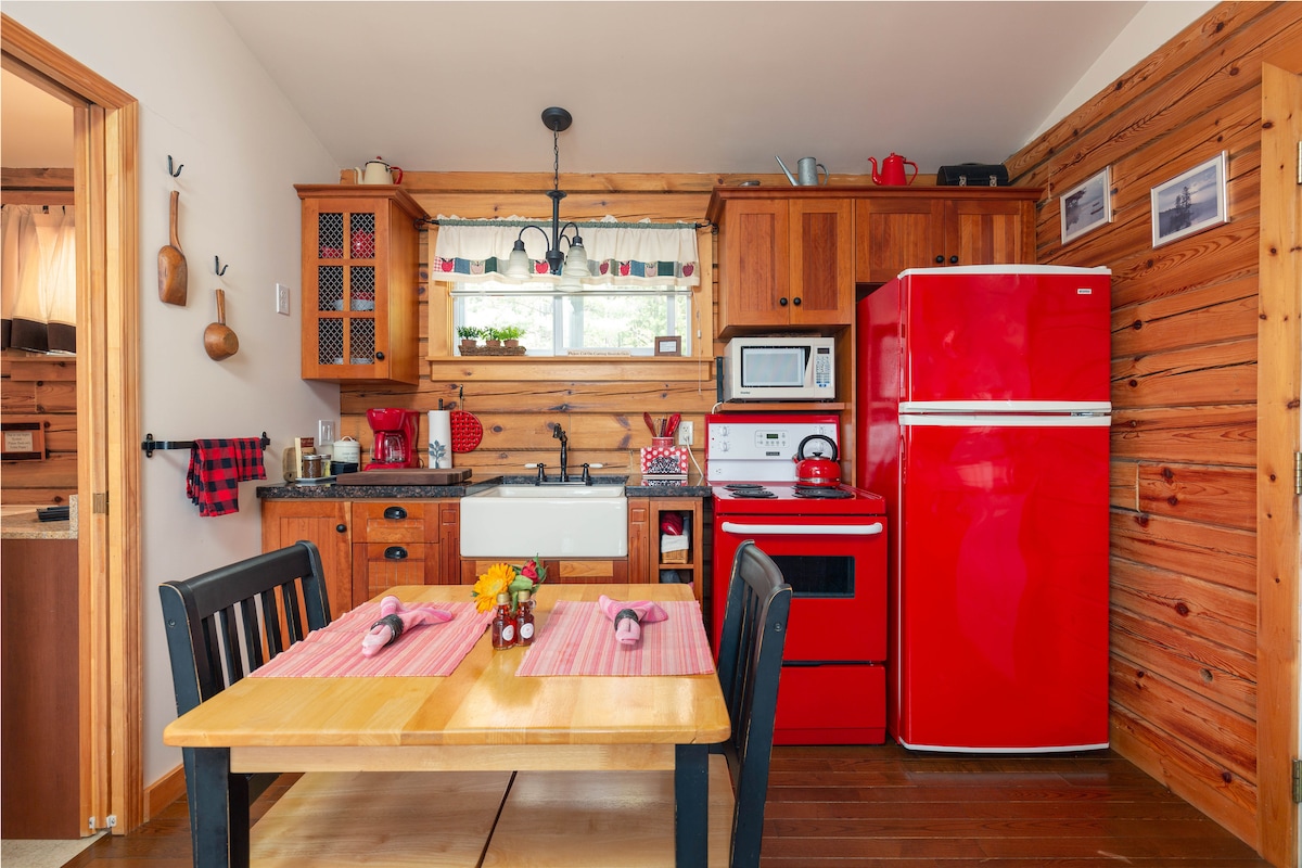 A compact kitchen features warm wooden walls with a mix of red accents and appliances, including a bright red refrigerator and stove. A wooden dining table is set with placemats and utensils, inviting guests to enjoy meals in a cozy atmosphere.
