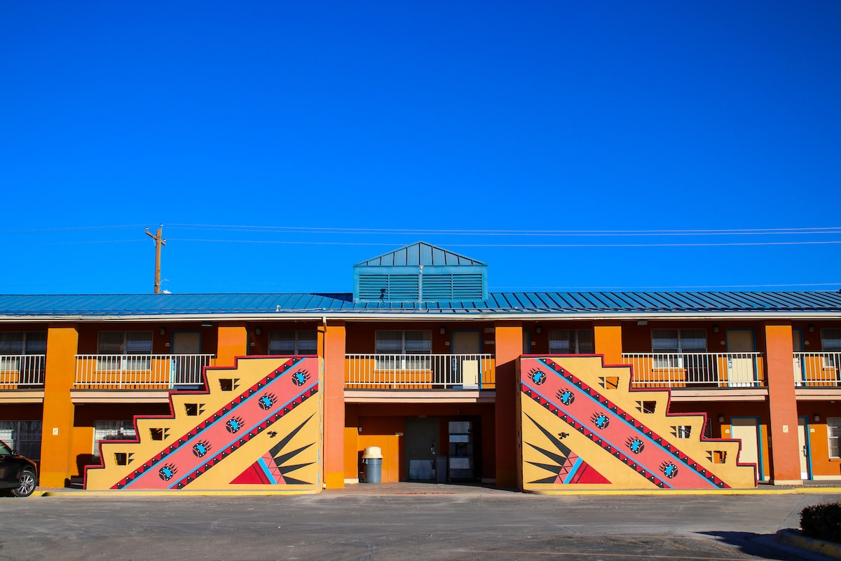 A colorful exterior of a two-story building is featured, displaying vibrant orange walls with artistic mural patterns. Balconies are visible on each level, and a modern glass structure is positioned at the top. A clear blue sky complements the vivid design.