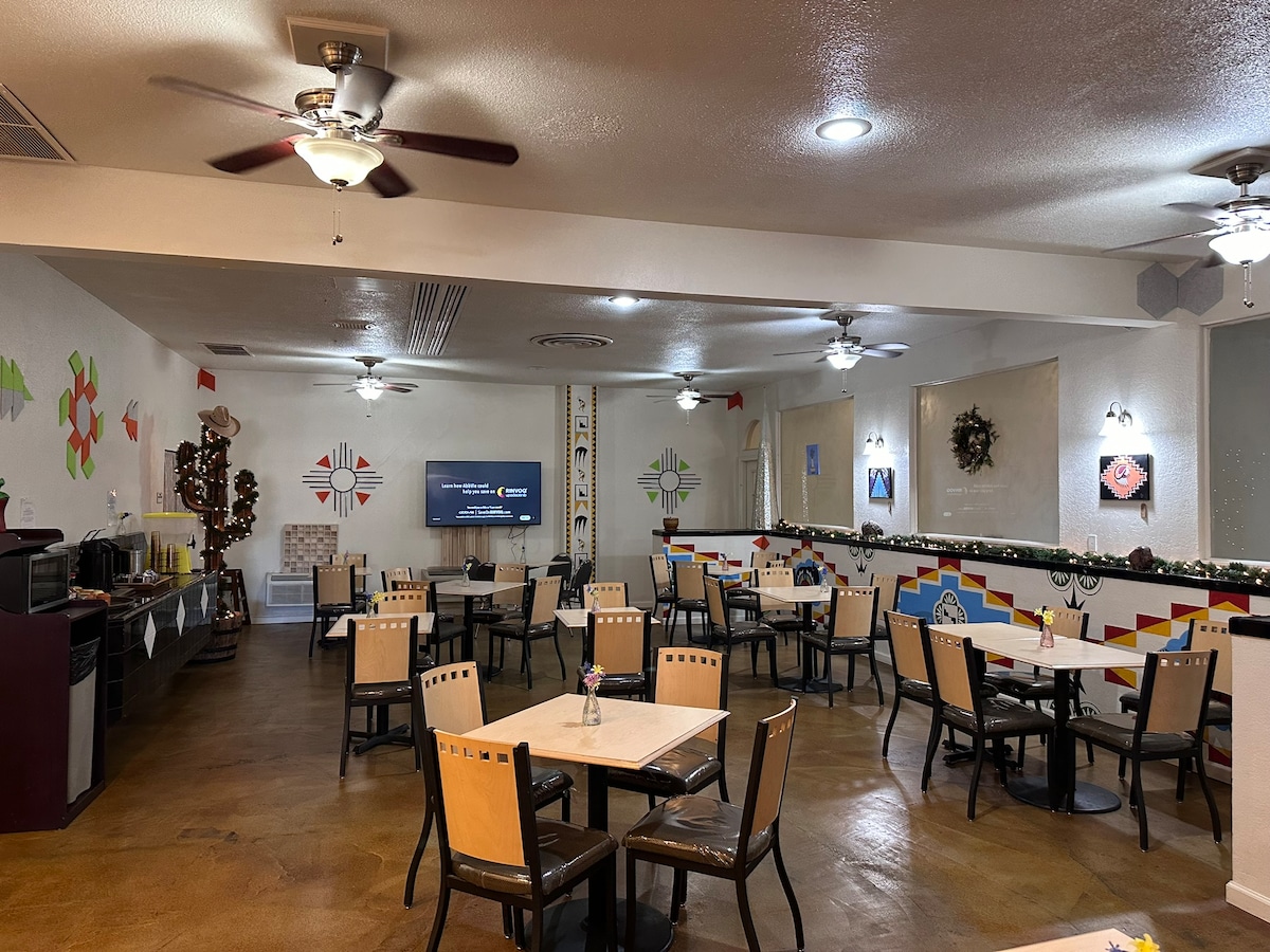 A dining area is arranged with several tables and chairs, featuring a warm, earthy floor. Walls are decorated with Southwestern motifs, and ceiling fans provide ventilation. A large television displays content, and small centerpieces are situated on each table, enhancing the communal dining experience.