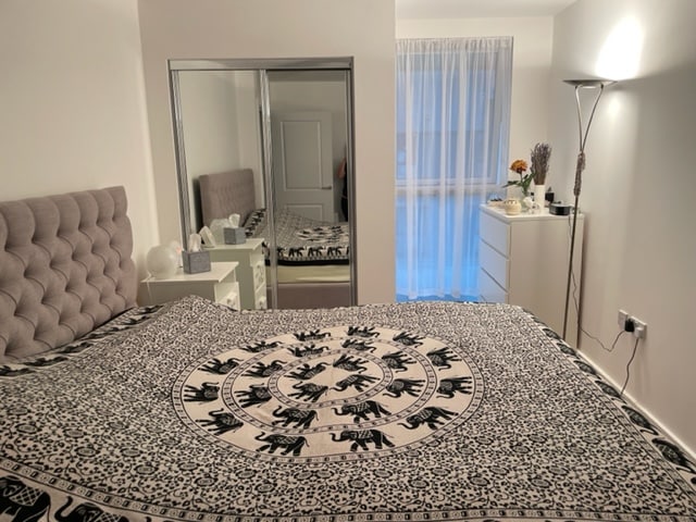 A well-arranged sleeping area is shown, featuring a spacious bed covered with an intricate patterned blanket. A large mirror reflects the room's light, while a dresser stands nearby, adorned with decorative items. Natural light filters through sheer curtains, enhancing the room’s brightness.