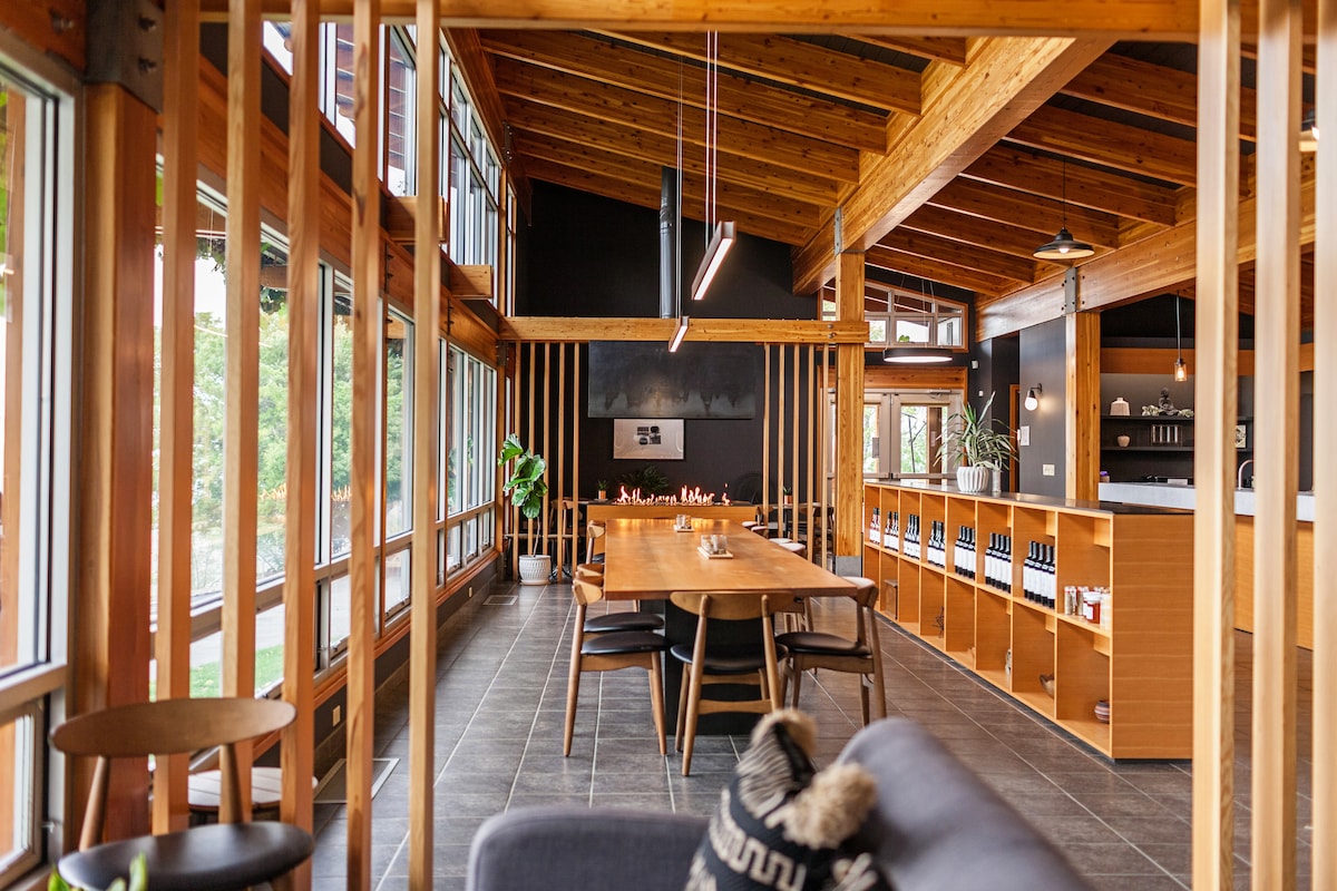 A spacious interior features large windows that bring in natural light, showcasing wooden beams and modern design. A long dining table is centered in the room, surrounded by chairs. Shelving displays wine bottles and plants, creating an inviting atmosphere.