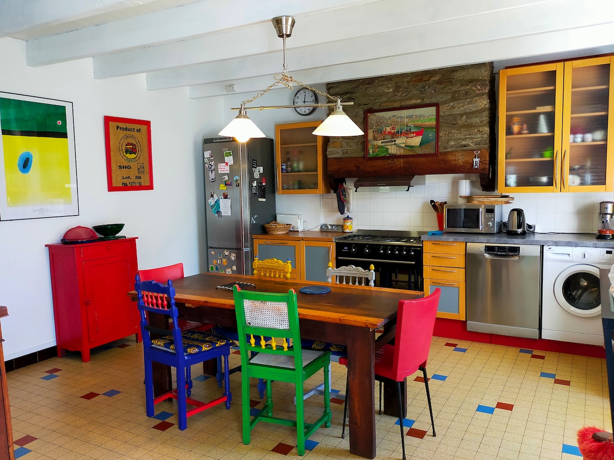 A well-equipped kitchen features modern appliances alongside rustic wooden elements. A large table with colorful chairs invites shared meals, while overhead light fixtures provide illumination. The decor mixes vibrant artwork and functional cabinetry, creating a welcoming space for cooking and gathering.