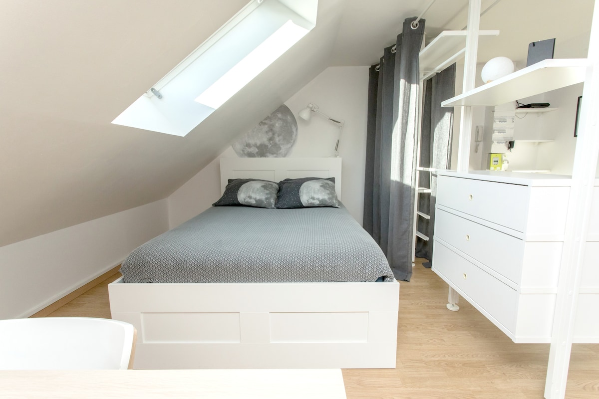 A cozy bedroom features a spacious bed dressed in grey linens, with soft pillows providing comfort. Natural light enters through a skylight, illuminating the room. A desk and shelving unit are positioned nearby, complemented by grey curtains that frame the window.