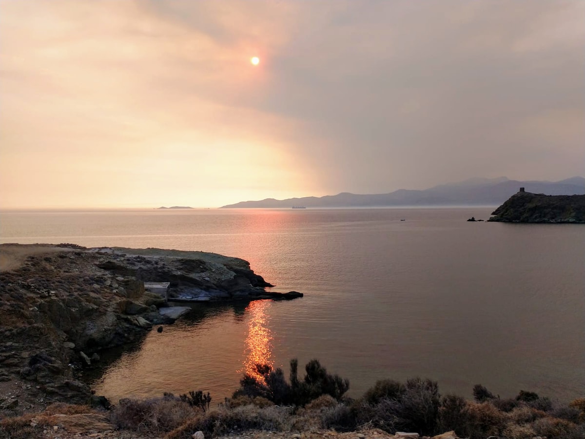 A serene seascape at sunset reveals soft orange and pink hues reflecting on calm waters. The shoreline features smooth rocky formations leading to a distant landmass visible across the sea. Gentle waves lap against the coast, contributing to the tranquil atmosphere.