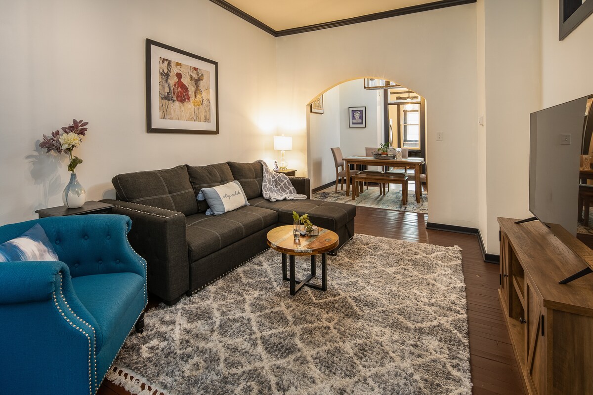 A comfortable living area is equipped with a dark sectional sofa and a vibrant blue armchair. A round coffee table sits atop a patterned area rug, while decorative floral art adorns the walls. A dining area is visible in the background, enhancing the open layout.
