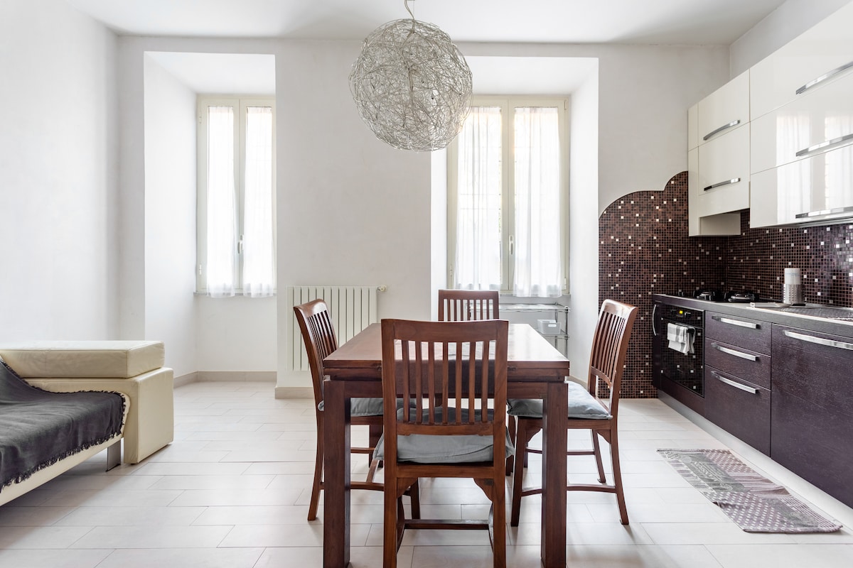 A dining area features a wooden table with four matching chairs, inviting a communal atmosphere. A modern light fixture hangs above the table. The kitchenette contains dark cabinetry, highlighted by a backsplash of shimmering tiles, while natural light fills the space through two windows.