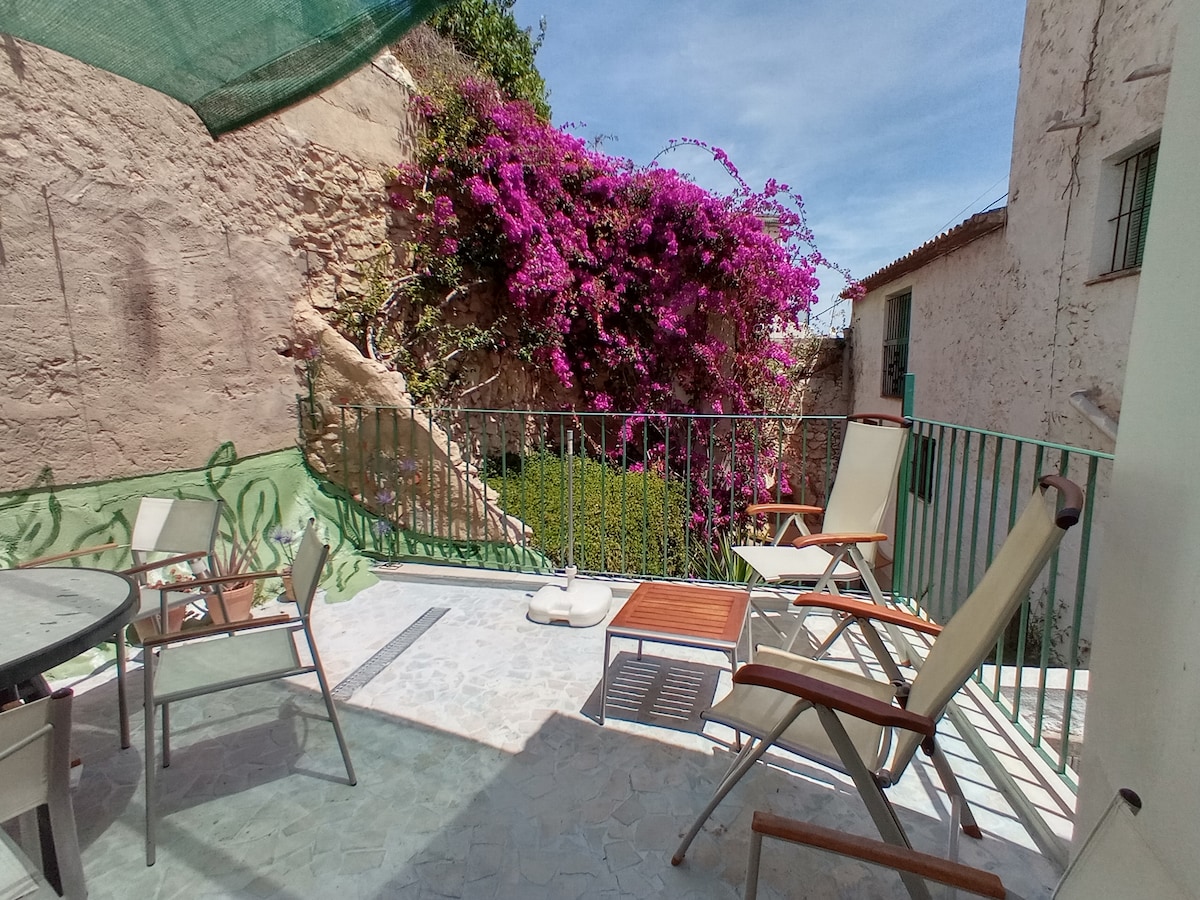 A spacious terrace displays a round table with four chairs, complemented by two lounge chairs. Lush purple flowers cascade over the surrounding wall, enhancing the serene outdoor setting. A small side table and a white cushion provide additional comfort.
