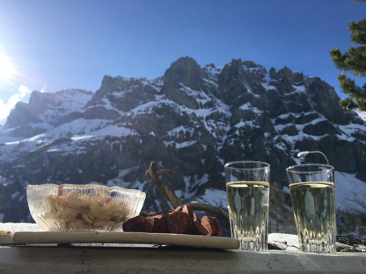 An outdoor setting displays a plate of sliced meat and a bowl of nuts alongside two glasses of light-colored beverage. The backdrop features snow-capped mountains under a clear blue sky, with sunlight creating a warm glow across the scene.