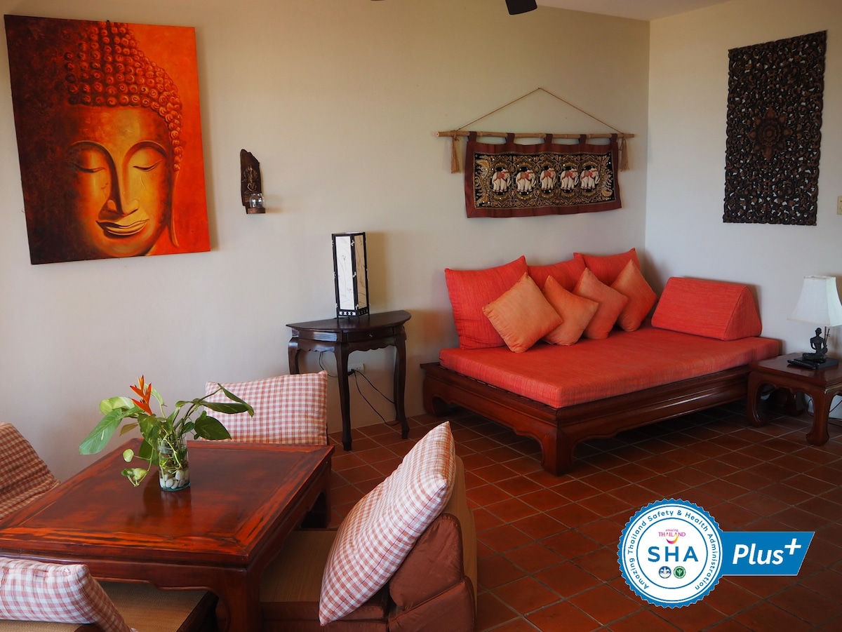A comfortable living space features warm, earthy tones with an inviting red seating area adorned with decorative cushions. A wooden table complements the relaxed atmosphere and is accompanied by two checkered chairs. Artistic wall hangings add a touch of cultural design to the room.
