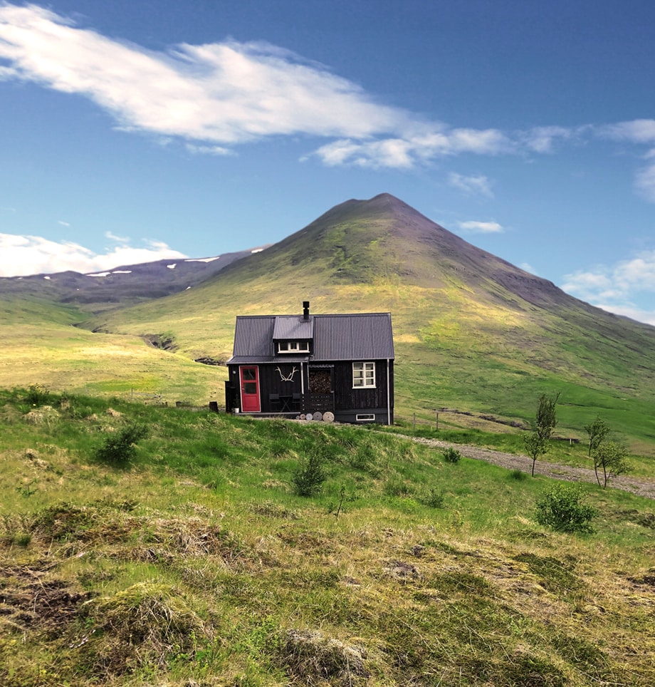 A charming cottage stands alone on a hillside, surrounded by lush green grass and gentle slopes. Majestic mountains rise in the background, their peaks partially covered in snow, under a bright blue sky with scattered clouds.