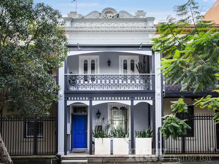  The image shows a classic two-story townhouse with intricate wrought-iron balconies and a bold, vibrant blue front door. The façade features ornate detailing along the roofline, highlighting its period architecture. Lush trees frame the house, adding a natural contrast to the sleek, grey-and-white exterior. Potted plants by the entrance and the upper balcony give the home a welcoming touch, while the overall design exudes elegance and charm in an urban setting.
