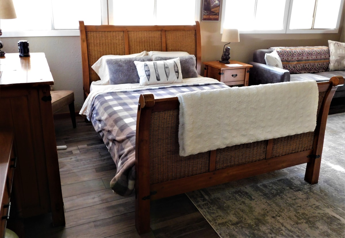 A cozy sleeping area features a wooden sleigh bed adorned with a patterned blanket. Soft pillows provide comfort, and natural light enters through large windows. Nearby furnishings include a rustic nightstand and a welcoming sofa, enhancing the warm atmosphere of the room.