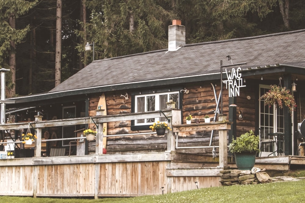 The exterior of the treehouse bed and breakfast is displayed, featuring a rustic wooden structure with a welcoming porch. Potted flowers adorn the railing, while a sign reading 'WAG TRAIL INN' adds charm. Surrounding trees are visible, enhancing the natural setting.