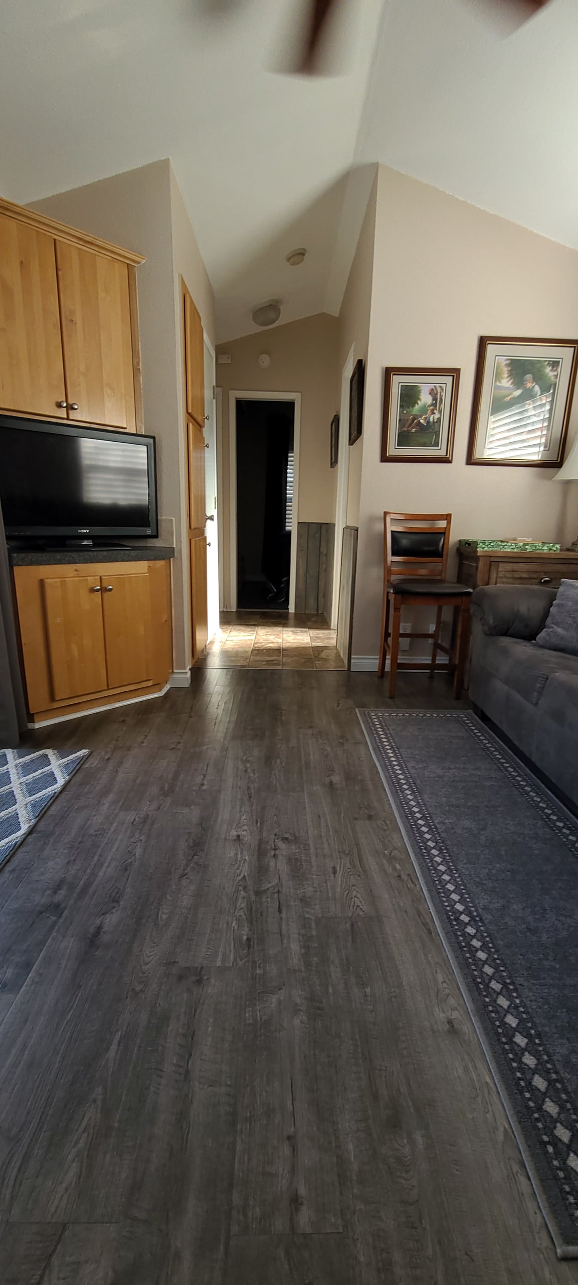 A narrow hallway is visible, featuring wooden cabinetry and a television mounted on the wall. A chair is positioned beside a small table, and the flooring is finished in a dark tone. Natural light illuminates the corridor, highlighting framed art pieces along the wall.