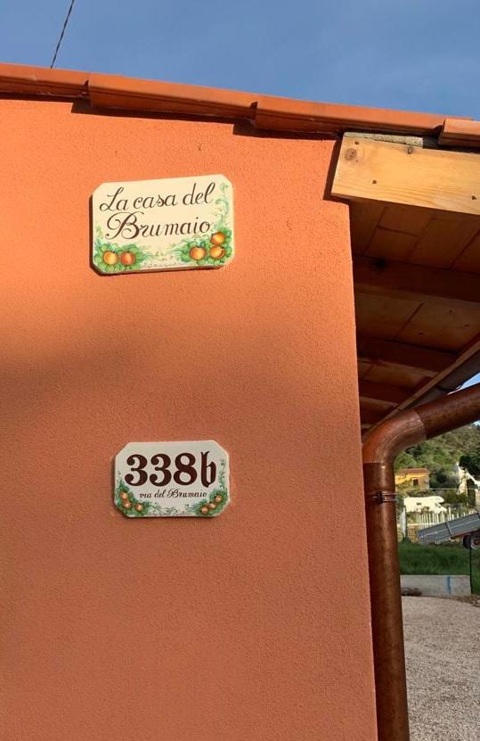 The exterior wall of a building presents two decorative plaques. The first plaque, titled 'La casa del Brumaio,' features artistic lettering and colorful designs. The second plaque indicates the address '338b,' framed with a simple white border. A portion of the roof eave is visible above.