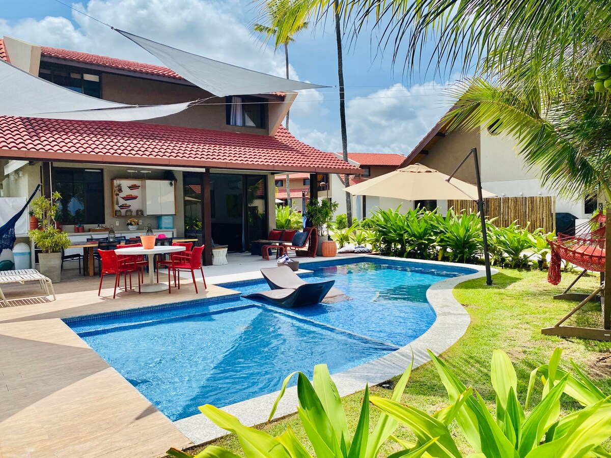 A private outdoor area features a clear blue swimming pool surrounded by greenery. Comfortable seating arrangements are positioned near the pool. A covered dining space with a table and chairs can be seen, offering a relaxed environment perfect for family gatherings.