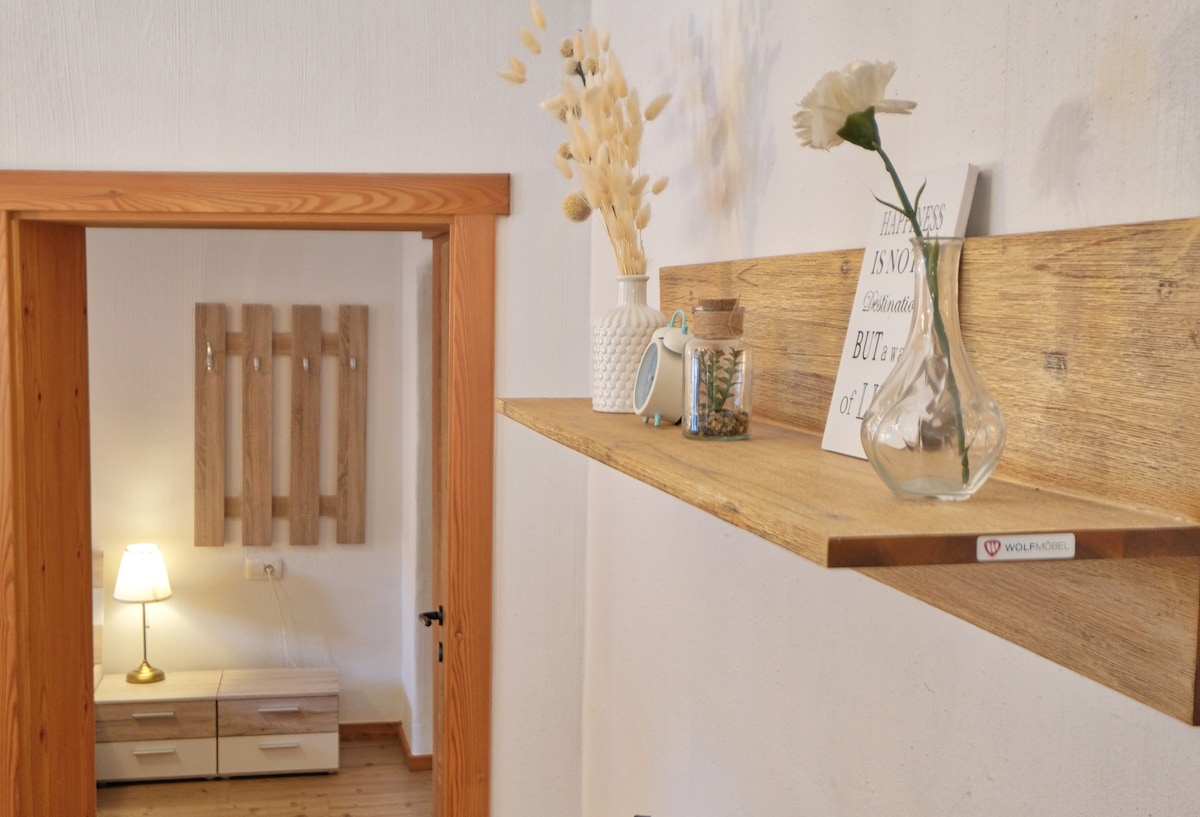A wooden shelf displays decorative items, including a vase with flowers, a small container, and a framed quote. The space features warm lighting from a nearby lamp, complemented by wooden accents in the door frame and furniture.