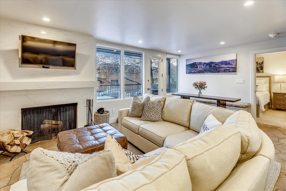 A spacious living area is presented with a wraparound couch and a flat-screen TV mounted above a stylish gas fireplace. Large windows allow natural light to fill the room, while a wooden table and decorative flowers add a refined touch.