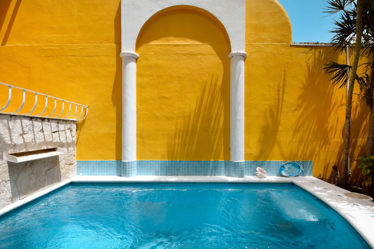 A vibrant yellow wall frames a dive pool, which is finished in azure tiles, reflecting the sunlight. A stone wall is visible to one side, and palm leaves provide a touch of greenery, enhancing the serene atmosphere of the pool area.