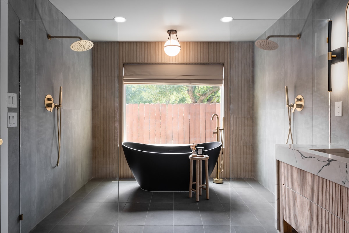 A contemporary bathroom features a freestanding black tub positioned beside a window with natural light. Two walk-in showers are equipped with elegant fixtures. A minimalist wooden stool and a marble countertop add to the clean, modern aesthetic.