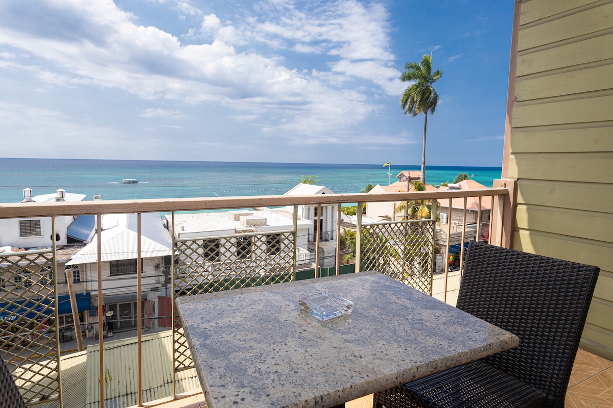 A private balcony offers a table with chairs, featuring a view of the Caribbean Sea. The horizon displays a blend of blue skies and water, with a palm tree standing in the foreground, creating a serene coastal setting.