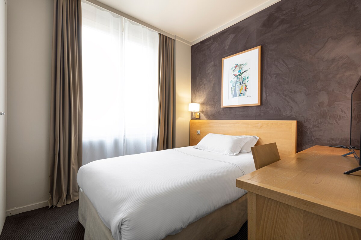 A cozy room features a comfortable bed with crisp white linens, accompanied by a wooden bedside table and a desk. Natural light enters through a large window covered with sheer curtains. A piece of artwork adds a touch of color to the textured wall.