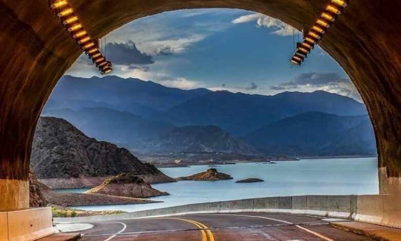 A view from within a tunnel frame reveals a winding road leading to a serene lake surrounded by mountains. The calm water reflects the sky and landscape, while warm overhead lights illuminate the tunnel entrance.