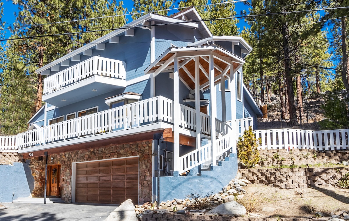 The exterior of the cabin showcases a two-story structure with a blue facade and stone accents. A spacious deck with white railings extends from the second floor. Tall pine trees surround the area, adding to the natural setting. A large garage door is visible at the base.