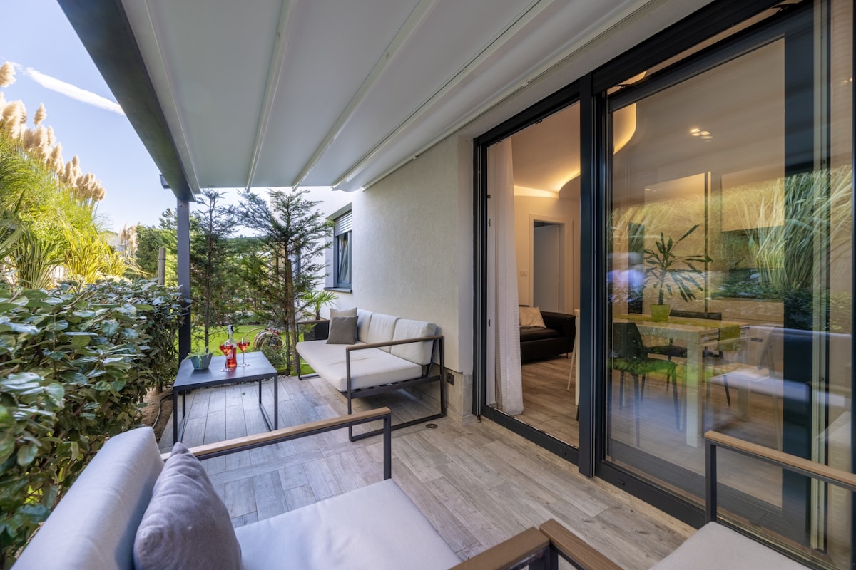 An outdoor patio area is furnished with comfortable seating and a small table. Lush greenery surrounds the space, creating a serene environment. Large glass doors lead to the interior, providing a connection between indoor and outdoor living.