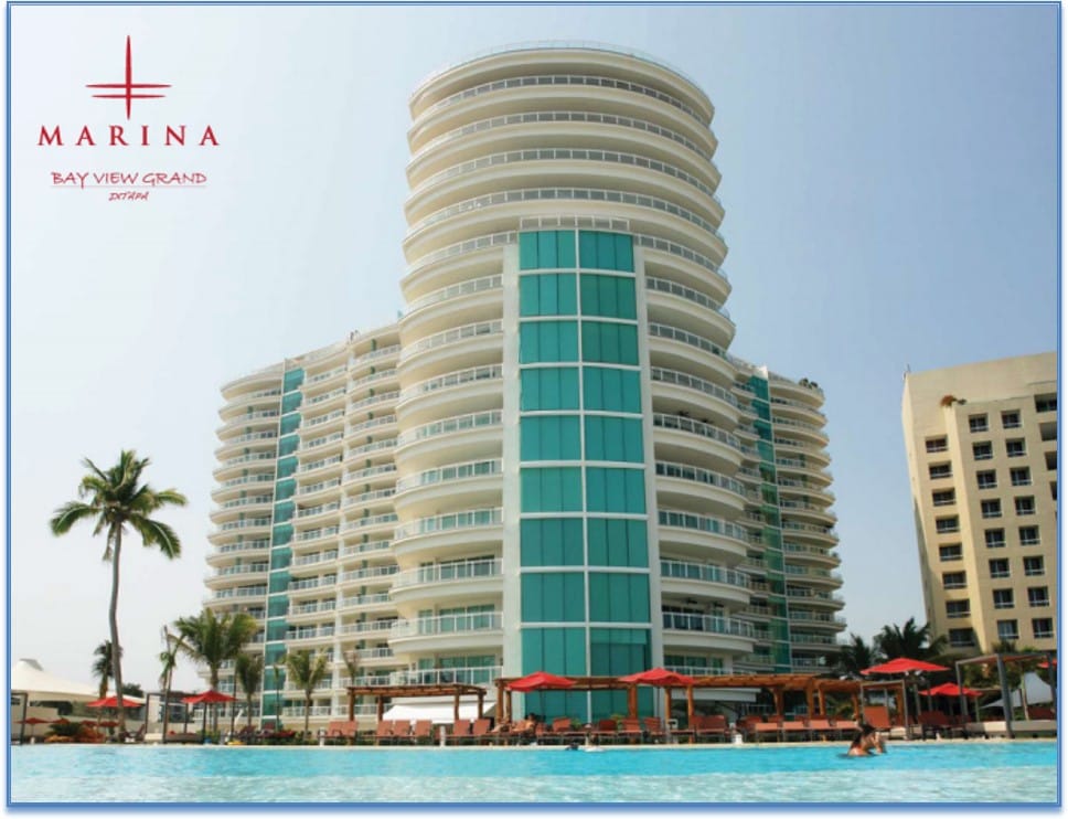 The image showcases a tall, modern condominium building with a rounded architectural design featuring large glass windows. Surrounding palm trees and lounge chairs are visible near a swimming pool, emphasizing a relaxed outdoor environment. The branding at the top indicates the location's name.