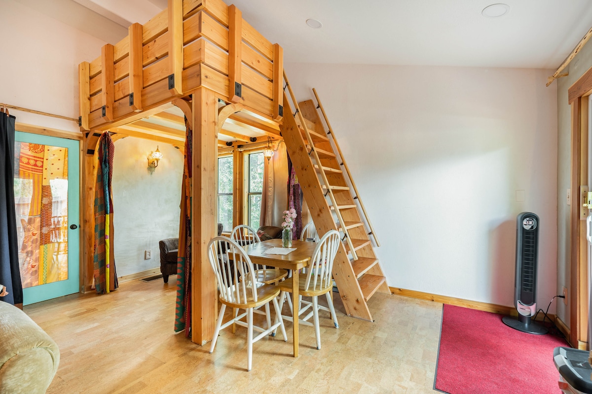 A spacious living area displays a wooden sleeping loft above, accessible by a sturdy ladder. Below, a round dining table with four white chairs invites gathering, while natural light brightens the room from a window. Soft furnishings create a cozy atmosphere, complemented by warm wood accents.