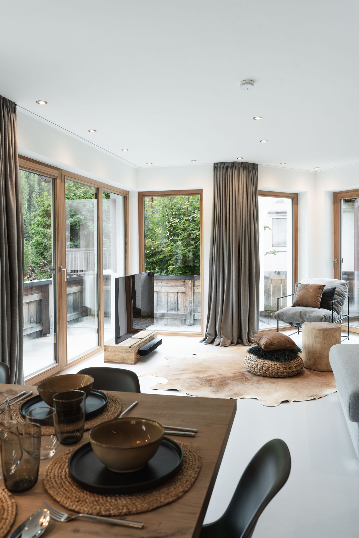 A modern dining area is displayed with a wooden table set for multiple guests. Light streams through large glass doors, illuminating the space. Natural materials, including a cowhide rug and woven seating, complement the room's contemporary design, while greenery is visible through the adjacent balcony.