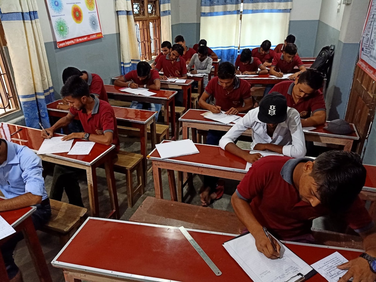 A classroom setting is depicted, showing students seated at wooden desks with red tabletops. The students are focused on writing, with papers and pens in front of them. Natural light filters through large windows adorned with light curtains, illuminating the room's calm atmosphere.