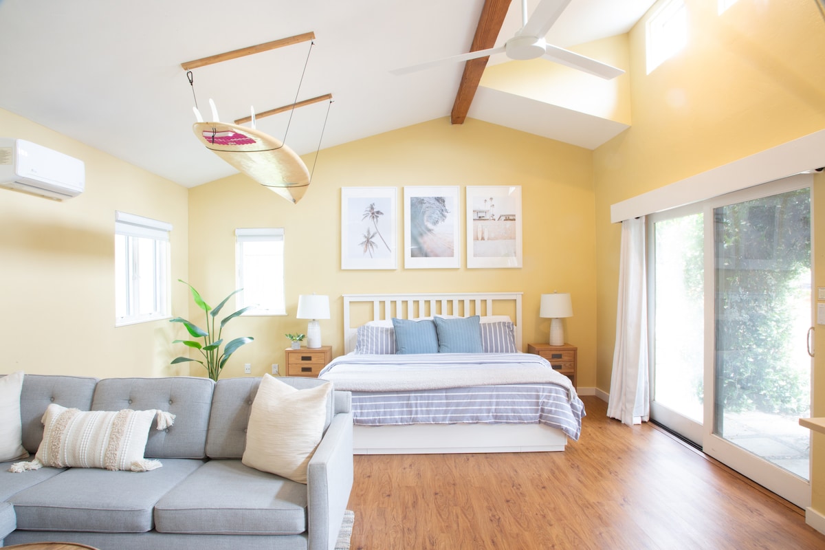 A bright and spacious bedroom features a king-sized bed with a light-colored duvet, complemented by bedside lamps on wooden tables. Large windows allow natural light to flood the room, and a comfortable sofa adds an inviting touch. A surfboard decoratively hangs from the ceiling.